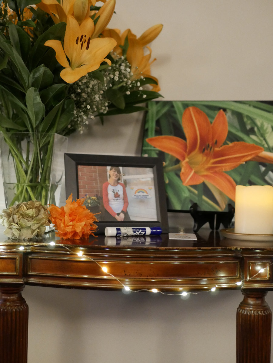 A memorial table with a photograph of a young woman with short brown hair and glasses. Behind her photo is a photograph of an orange lily that she had taken. Real lillies stand in a vase on the other side of the table. A name tag and dry erase marker can also be seen on the table, along with orange and gold paper flowers. 
